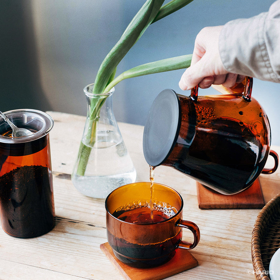 Person pouring tea from an amber-colored coffee server into a matching cup on a wooden table.