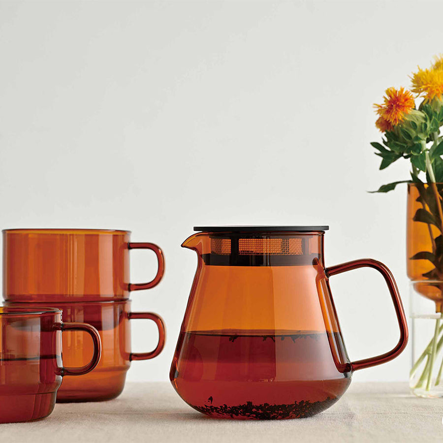 Amber-colored glass coffee server and mugs on a light surface with a vase of flowers in the background.
