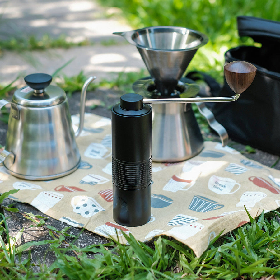Outdoor coffee brewing setup featuring Hario Kabuto manual grinder with wooden handle, stainless steel gooseneck kettle, and metal pour-over dripper on a patterned cloth – ideal for coffee lovers on the go.