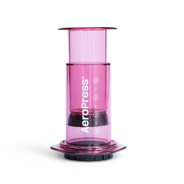 A pink AeroPress coffee maker made of shatterproof Tritan material, featuring a clear chamber, plunger, filter cap, and seal.