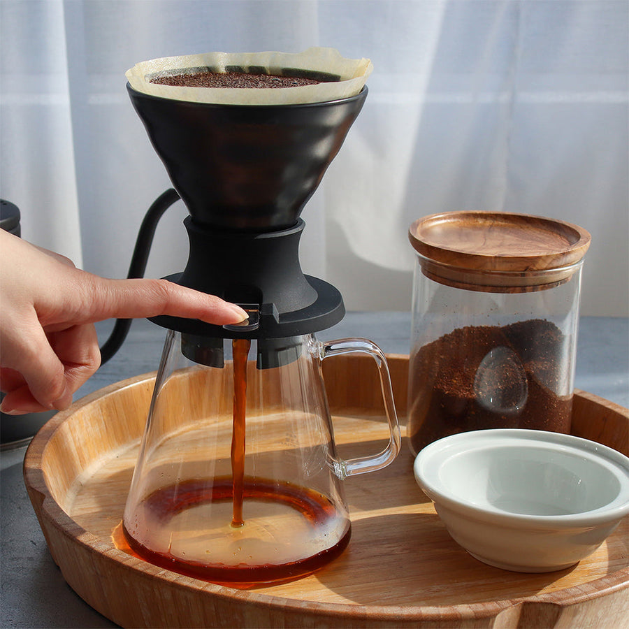 Hand pouring coffee into a glass carafe with a wooden tray and Hario Switch V60 Immersion Dripper