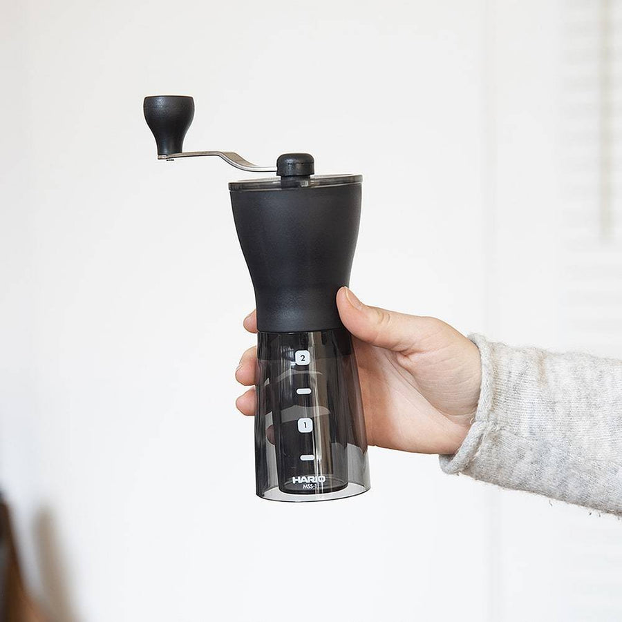 Hand holding the Hario Mini Mill PLUS manual coffee grinder, showing black top, crank handle, and transparent base with 1 and 2 cup measurement markings.