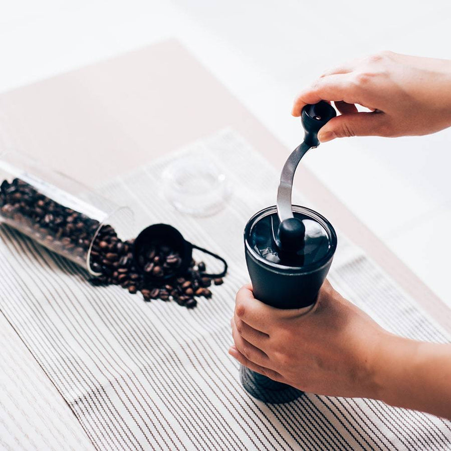 Person grinding coffee beans with a Hario Mini Mill PLUS hand grinder, surrounded by spilled beans, a scoop, and a striped cloth surface.