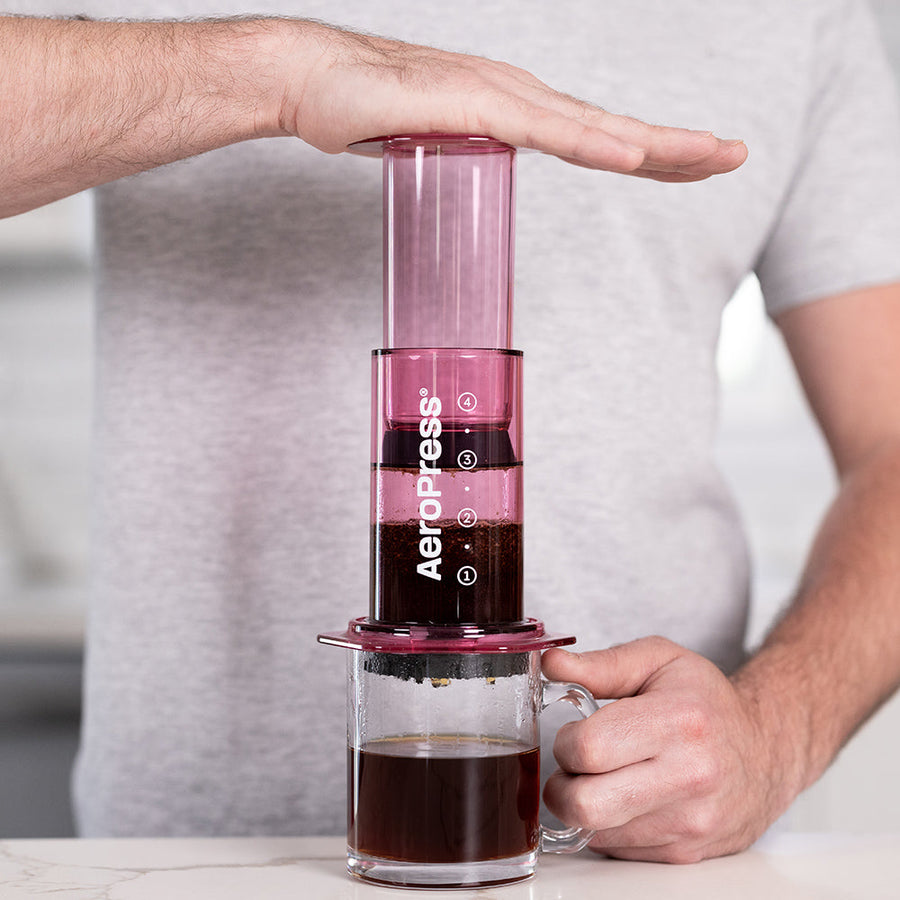A person plunging a pink AeroPress coffee maker made of shatterproof Tritan material, featuring a clear chamber, plunger, filter cap, and seal.