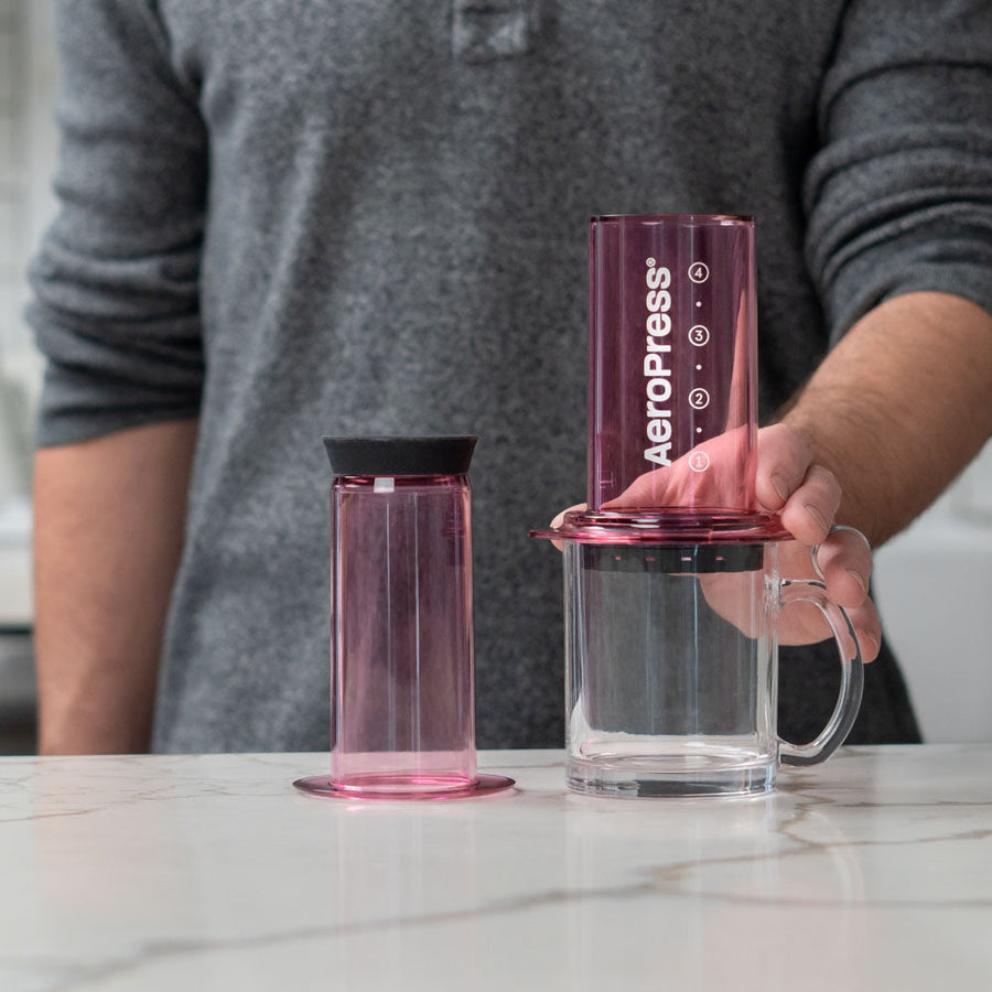 A pink AeroPress coffee maker made of shatterproof Tritan material, featuring a clear chamber, plunger, filter cap, and seal.