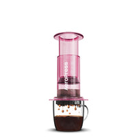A pink AeroPress coffee maker made of shatterproof Tritan material, featuring a clear chamber, plunger, filter cap, and seal.