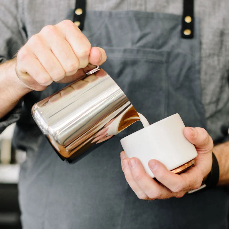 Fellow Eddy Steaming Pitcher - Polished Steel 18oz pouring milk into a coffee cup in the hands of a barista