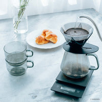 Coffee brewing process on a kitchen counter with Hario coffee maker, cups, and pastries.