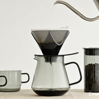 Coffee brewing process with a clear glass carafe and black coffee filter on a light background.