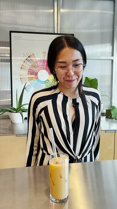 A woman in a black and white striped top looking at an iced latte in a tall glass on a metal counter.
