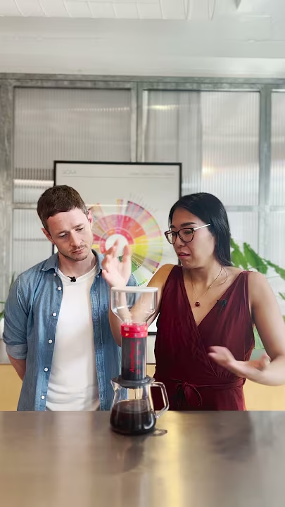A man in a denim shirt and white t-shirt and a woman in a red dress brew coffee on a stainless steel counter.