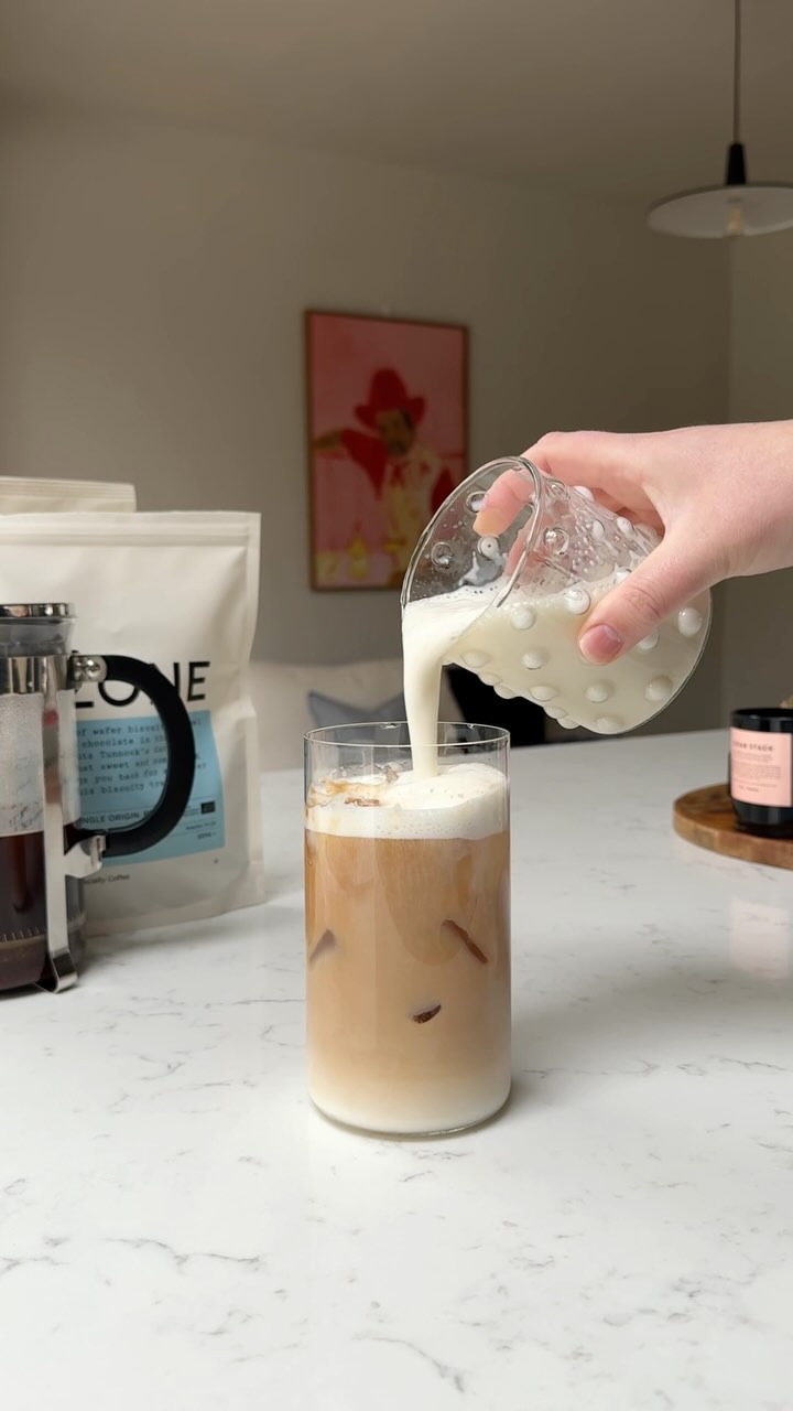 Foamed cold milk being poured on top of an iced latte in a tall glass on a kitchen counter in a modern home.