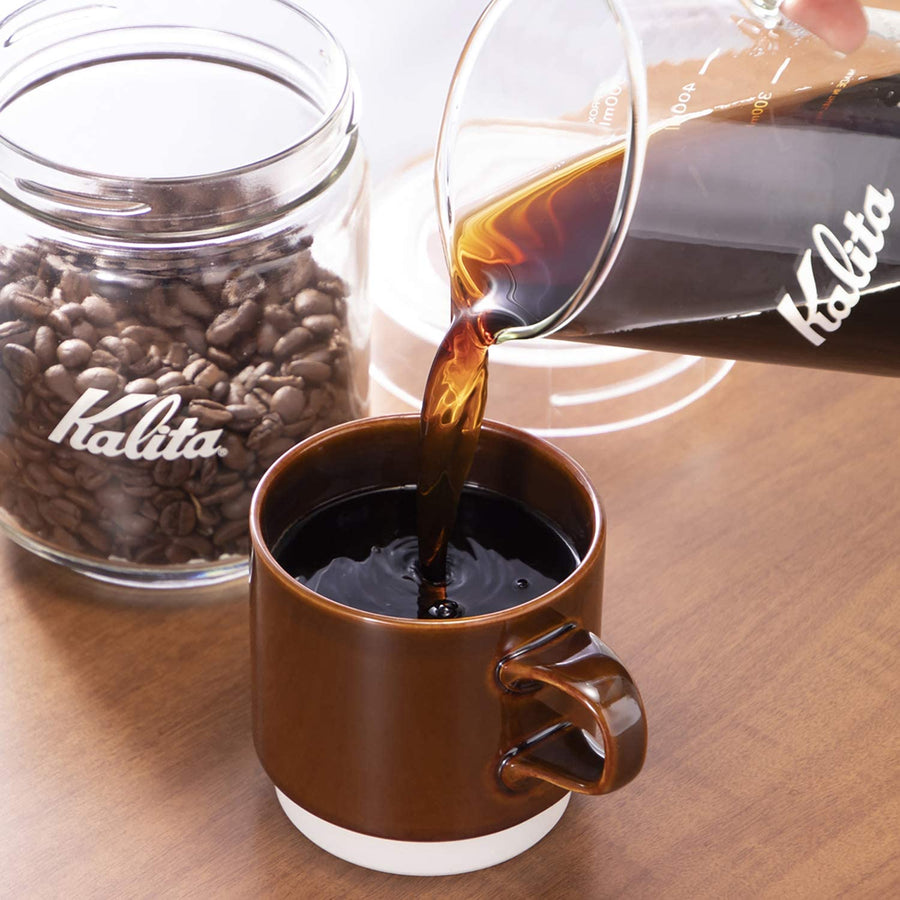 Kalita Jug Glass Server - Jug 500 (500ml) lifestyle image showing jug 500 server being used to pour coffee into a brown cup with a glass jar of coffee beans in the background